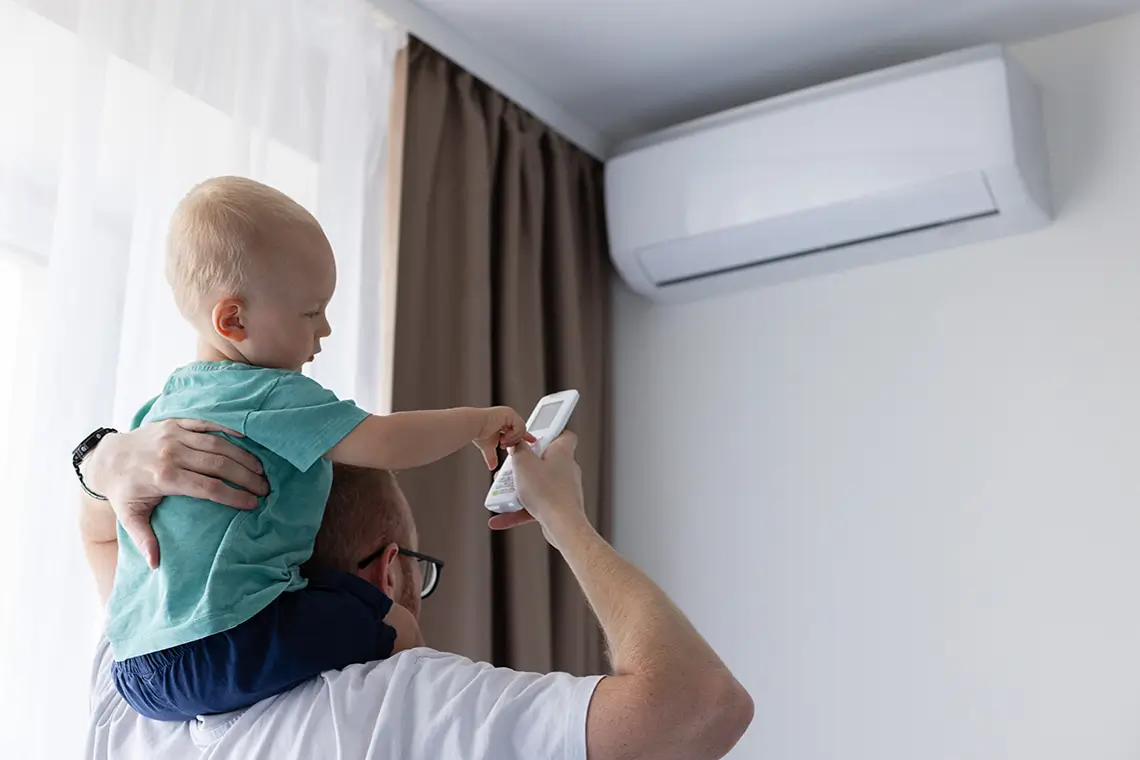 Vater hält Kind auf den Schultern, das mit einer Fernbedienung eine Klimaanlage im Zimmer bedient Vater hält Kind auf den Schultern, das mit einer Fernbedienung eine Klimaanlage im Zimmer bedient