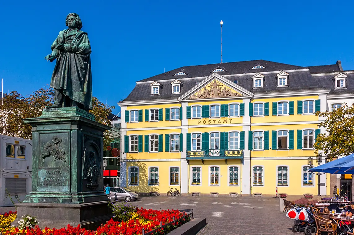 Historisches gelbes Postamt in Bonn mit Beethoven-Denkmal auf dem Marktplatz bei blauem Himmel.